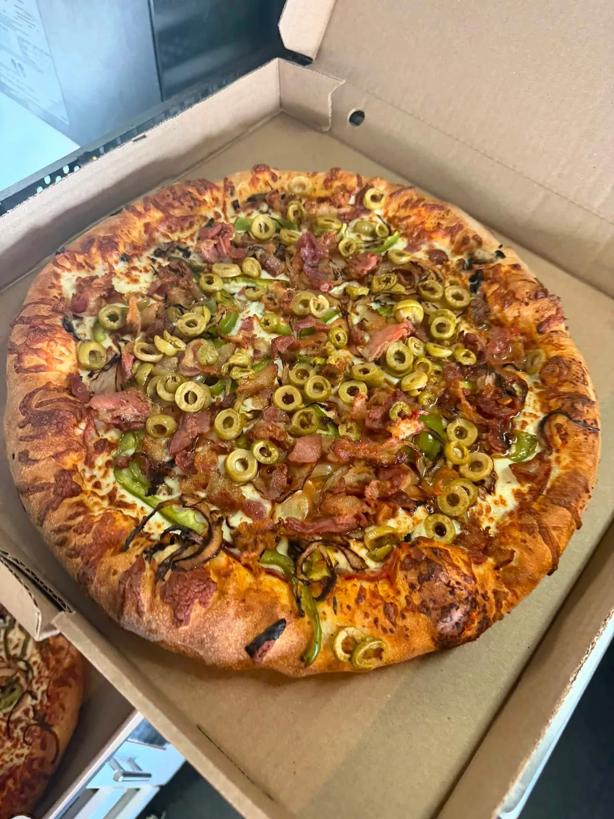Pizza garnie de bacon, de viande fumée, d’olives vertes et de piments verts, dans sa boîte.