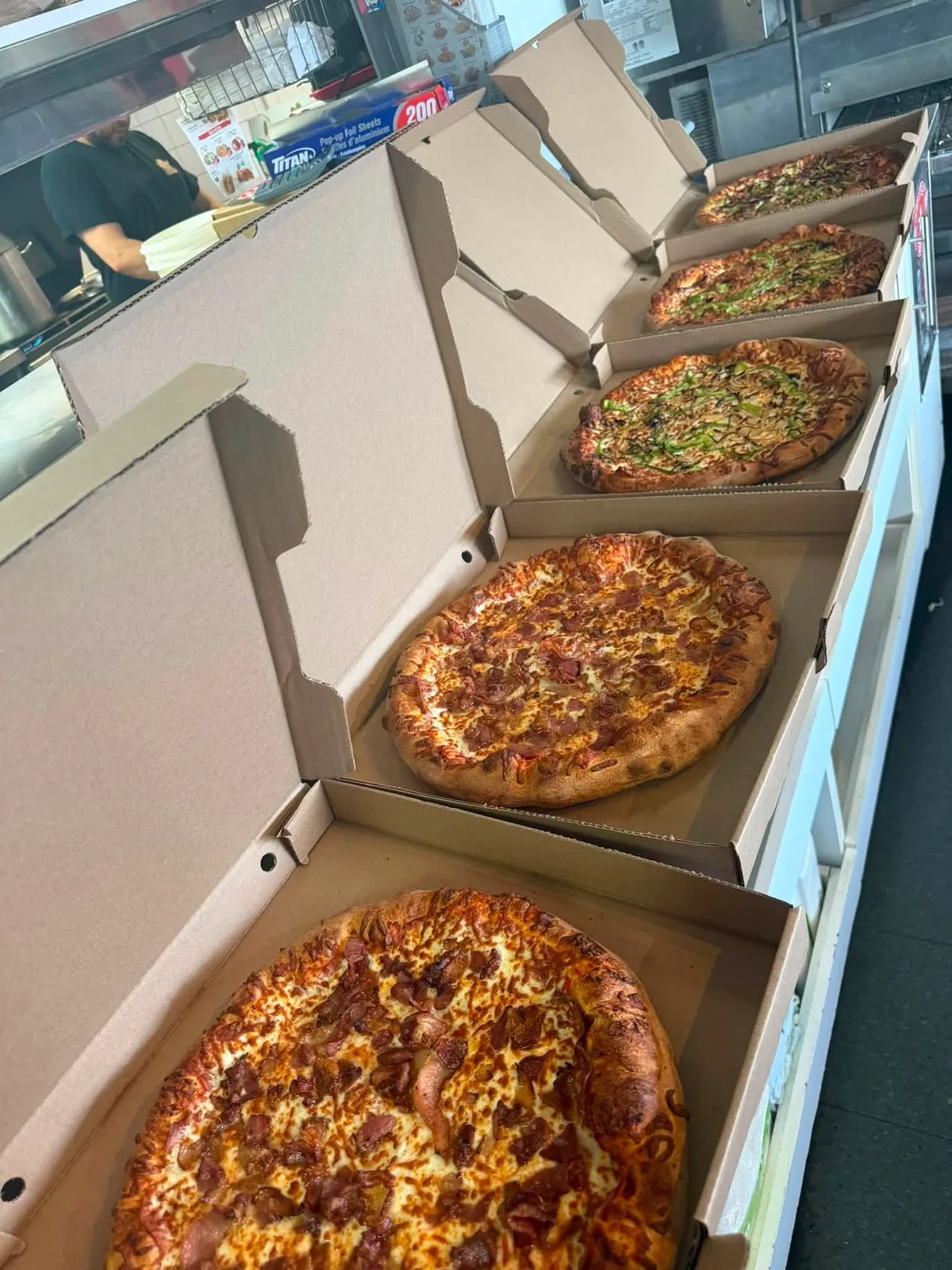 Plusieurs pizzas dans leurs boîtes ouvertes, alignées sur le comptoir de la cuisine du restaurant.